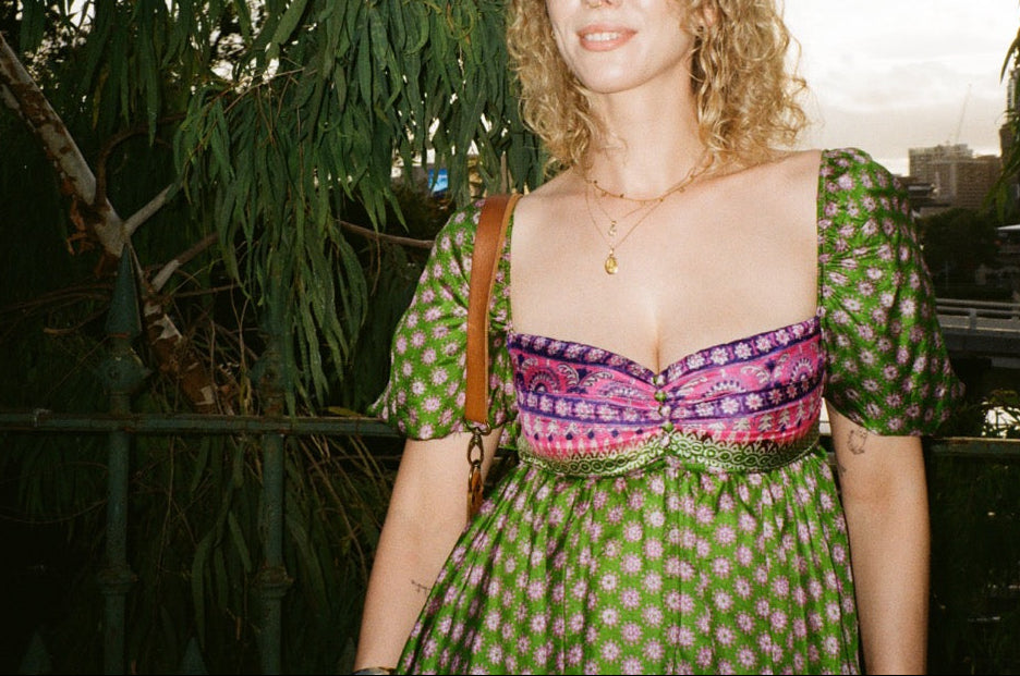 Woman in a green patterned dress with a colorful top standing outdoors among trees.